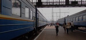Lvivin juna-asema vuonna 2012. Siniset matkustajajunat seisovat asemalaitureilla, ja ihmiset liikkuvat laiturilla, osa kävellen kohti junia ja osa poispäin. Yläpuolella kulkee metallinen kävelysilta, ja taustalla näkyy sähkölinjoja ja asemarakenteita.