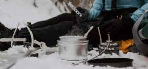 Henkilö istuu lumisessa metsässä talvivarusteissa ja keittää ruokaa retkikeittimellä. Höyryävä kattila on Trangia-merkkinen, ja vieressä näkyy Primus-kaasupoltin. Maassa on myös lumikengät ja muita retkeilyvarusteita.