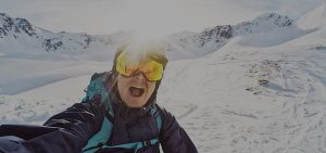 Hiihtovaeltaja ottaa innostuneen selfien lumisessa vuoristomaisemassa. Hänellä on laskettelulasit, kypärä ja talvivarusteet, ja aurinko paistaa suoraan takaa luoden heijastuksen laseihin. Taustalla avautuu lumipeitteisiä vuoria ja laskujälkiä rinteessä.