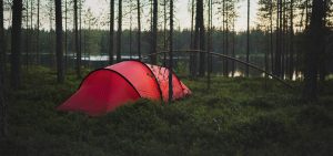 Punainen tunneliteltta on pystytetty vihreälle varvikolle rauhallisen metsälampimaiseman äärelle. Teltta on ympäröity hoikilla mäntyjen rungoilla, ja ilta-aurinko siivilöityy puiden välistä heijastuen veden pinnasta.