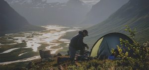 Retkeilijä pystyttää leiriä vuoristoisessa maisemassa ilta-auringon valossa. Edessä avautuu jylhä näkymä mutkitteleviin jokiin ja laaksoon. Tumma kupoliteltta seisoo rinteellä, ja retkeilijä järjestelee varusteitaan.