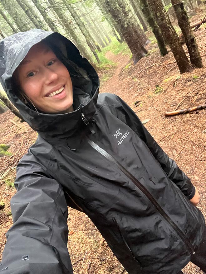 Amanda hymyilee selfie-kuvassa metsässä päällään musta Arc'teryx kuoritakki.