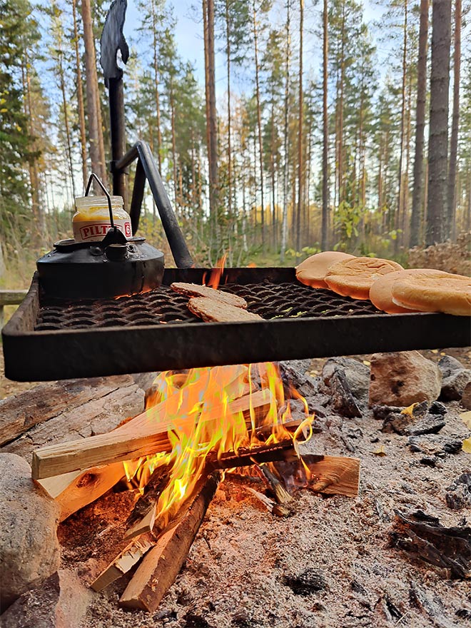 Laavupaikan nuotiolla eväät lämpiämässä. Piltti-purkki Trangian kahvipannussa vesihauteessa ja lihapiirakat ritilällä.