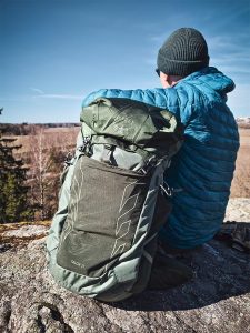 Henkilö sinisessä takissa ja vihreä Osprey Talon 33 -reppu selässä istuu kalliolla maisemia katsellen.
