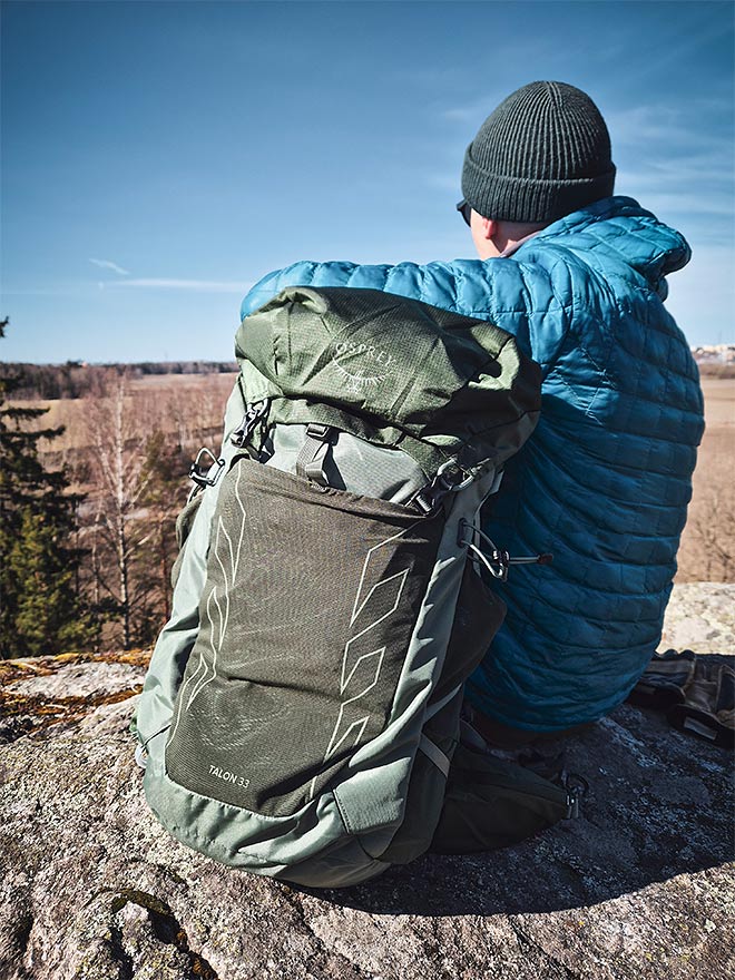 Henkilö sinisessä takissa ja vihreä Osprey Talon 33 -reppu selässä istuu kalliolla maisemia katsellen.