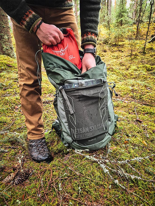 Henkilö pakkaa punaista pakkauksta Osprey Talon 33 -reppuun metsäisessä maastossa.