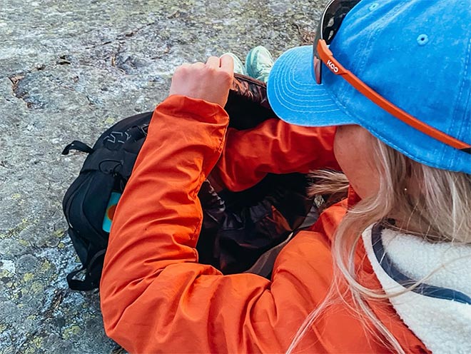 Retkeilijä, jolla on sininen lippis ja oranssi takki, kaivaa tavaroita repustaan taukopaikalla kallion päällä.