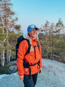 Retkeilijä seisoo hymyillen kallioisella näköalapaikalla, yllään oranssi takki ja sininen lippis.