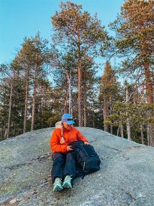 Retkeilijä istuu kallion päällä metsämaisemassa ja selaa mustaa reppuaan iltavalossa.