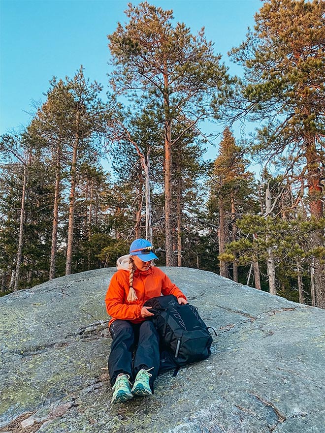Retkeilijä istuu kallion päällä metsämaisemassa ja selaa mustaa reppuaan iltavalossa.