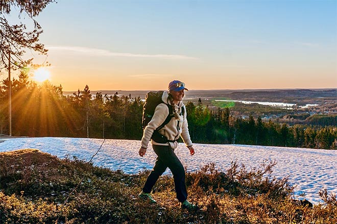 Retkeilijä vaeltaa iloisena ilta-auringossa lumilaikkujen peittämällä rinteellä, taustalla avautuu laaja metsä- ja järvimaisema.