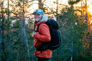 Hymyilevä retkeilijä nauttii ilta-auringosta metsäisessä maisemassa, yllään lämmin takki ja selässään musta reppu.