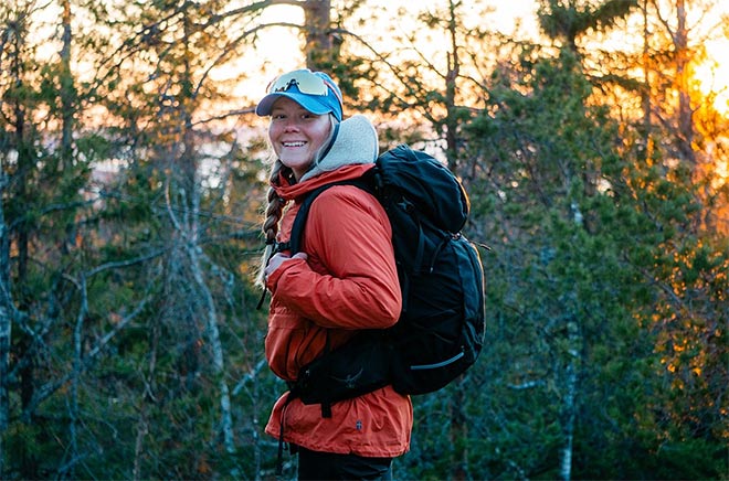 Hymyilevä retkeilijä nauttii ilta-auringosta metsäisessä maisemassa, yllään lämmin takki ja selässään musta reppu.
