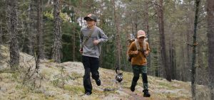 Kaksi lasta ja pentukoira kävelevät sammaleisella metsäpolulla tiheiden kuusten keskellä; vasemmalla lapsi harmaassa fleecepaidassa ja vihreässä lippalakissa, oikealla lapsi okrankeltaisessa fleecetakissa ja beigen värisessä lippalakissa, molemmilla reppu selässä ja niiden välissä nuori husky-pentu.
