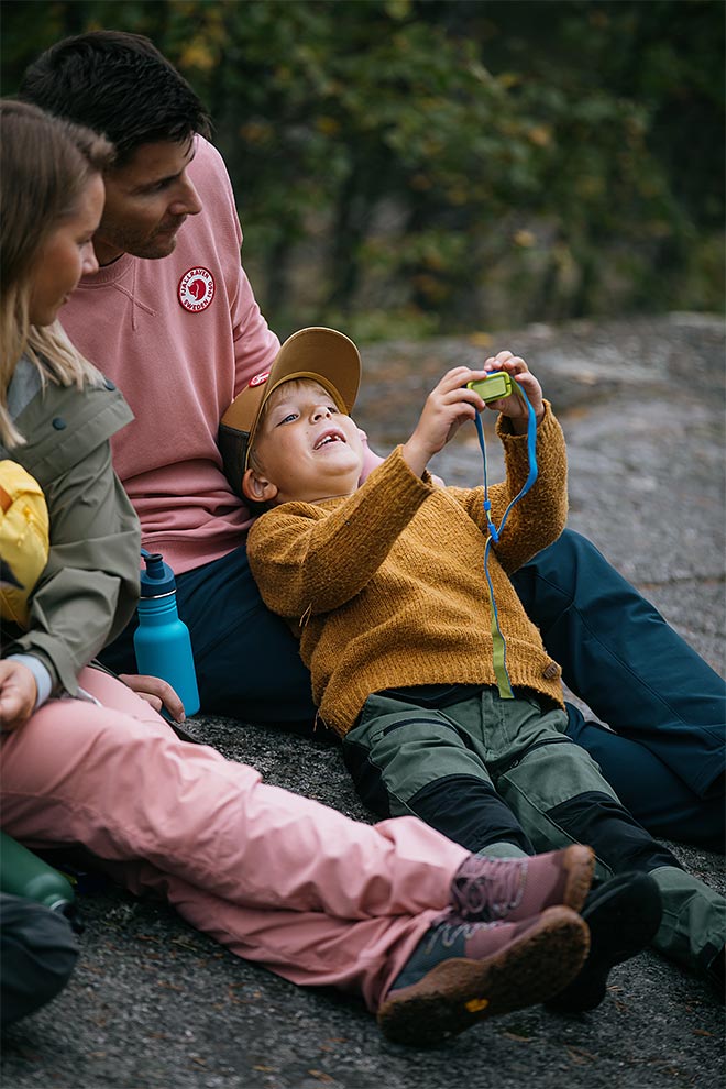 Perhe istuu kallioisella penkereellä metsässä; lapsi makaa aikuisen sylissä ja kohottaa kiikaria kohti taivasta, vanhemmat istuvat vieressä.