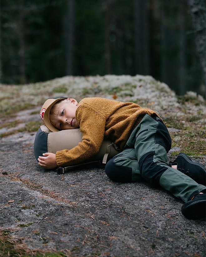 Lapsi oranssiokran värisessä fleecetakissa makaa kallioisella maastolla kylki ja päälevy repaselkäntyynynä, ympärillä sammalpeitteistä kalliota ja hentoja kuusenrunkoja.