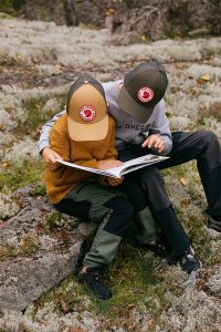 Kaksi lasta istuu sammaleisella kalliolla metsässä, molemmilla lippalakki päässä, ja lukee yhdessä avattua kirjaa.