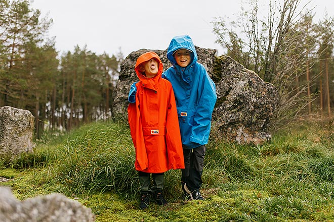 Kaksi lasta seisoo sammaleisella ja kivikkoisella metsämaastolla; vasemmalla lapsi oranssinpunaisessa sadetakissa, oikealla lapsi kirkkaan sinisessä sadetakissa, molemmilla huput päässä ja taustalla havupuita sekä kalliota.