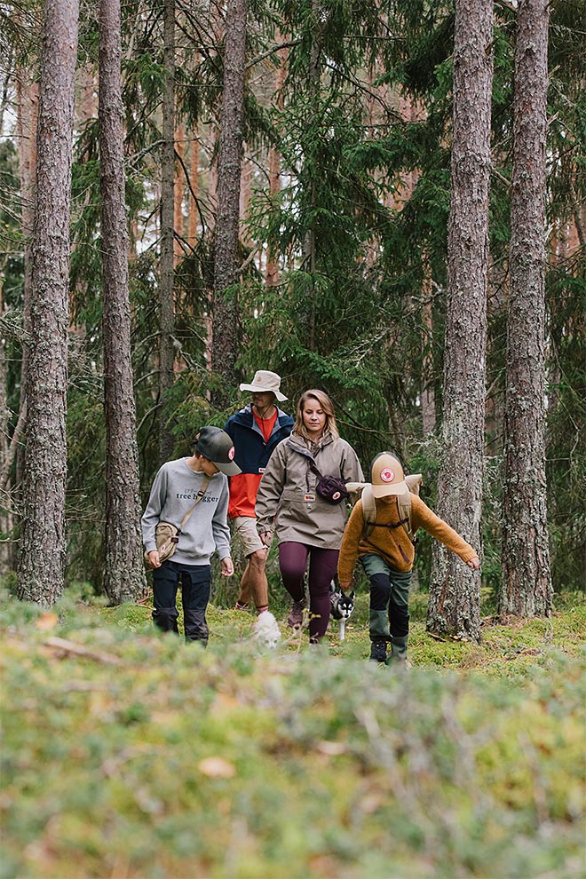 Perhe kävelee metsäpolulla sammaleen ja kuusien keskellä; etummaisena lapsi okrankeltaisessa fleecetakissa ja lippalakissa, vieressä toinen lapsi harmaassa fleecetakissa ja retiisilakkipäisessä lippalakissa, takana nainen vaaleanruskeassa takissa ja mies oranssinkirjavatakki päällään, kaikki kulkevat polkua pitkin tiheässä kuusikossa.