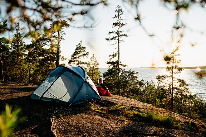Sininen retkiteltta on pystytetty kallioiselle niemekkeelle järven rannalla, ympärillä mäntyjä ja horisontissa pehmeä auringonlaskun valo siivilöityy puiden lomasta.