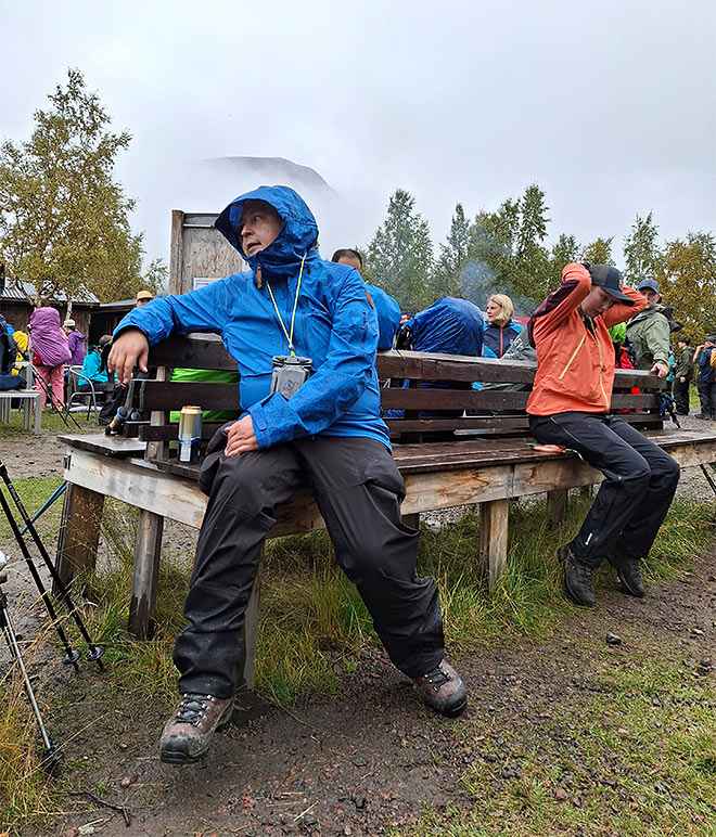 Sadeasuihin pukeutuneita vaeltajia istuu penkillä taukopaikalla metsässä.