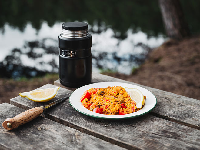 Musta ruokatermospullo puisella pöydällä. Vieressä valkoisella lautasella risotto.