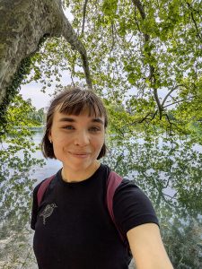 Retkeilijä ottaa selfien lehtipuun alla järven rannalla.