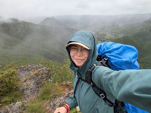 Retkeilijä ottaa selfien sateisella vuorenrinteellä reppu selässään, sumuinen laakso taustalla.
