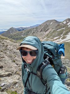 Retkeilijä ottaa selfien aurinkolasit päässä ja suuri reppu selässään lumilaikkuisten vuorten keskellä.
