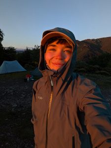 Retkeilijä ottaa selfien telttojen vieressä auringonlaskun valaistessa kasvot ja metsäisen vuoristomaiseman.