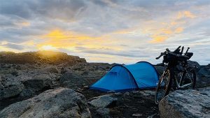 sininen teltta ja bikepacking-pyörä kivikkoisella leiripaikalla auringon laskiessa.