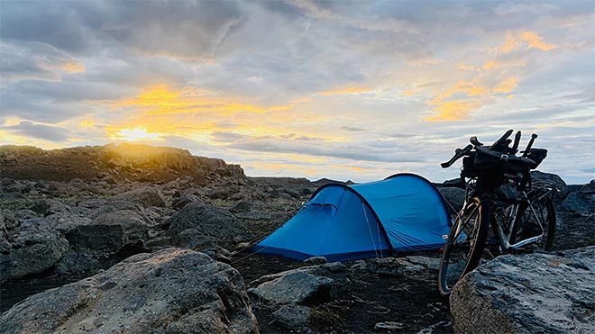 sininen teltta ja bikepacking-pyörä kivikkoisella leiripaikalla auringon laskiessa.