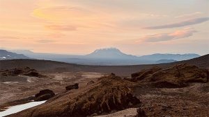 lumilaikkuinen vuori kohoaa laajan vulkaanisen ylänkömaiseman takana auringonlaskussa.