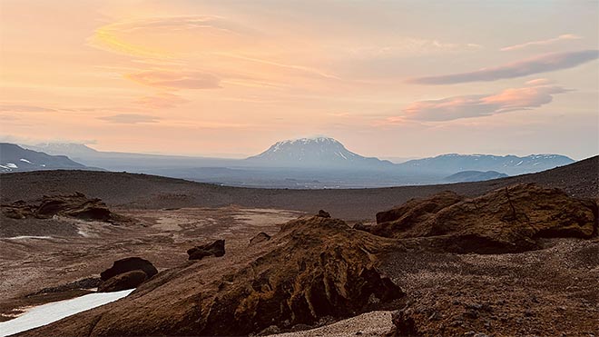 lumilaikkuinen vuori kohoaa laajan vulkaanisen ylänkömaiseman takana auringonlaskussa.